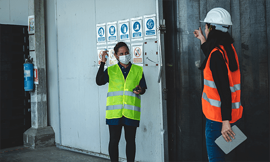 Photo of two workers wearing PPE standing in front of a warehouse wall featuring several safety signs.