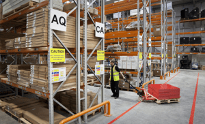 Photo of a man pulling a trolley between two warehouse aisles marked with aisle markers and racking load notices.