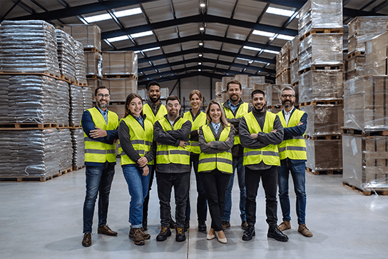 A team of warehouse employees stands and smiles at the camera inside a large well-lit warehouse.
